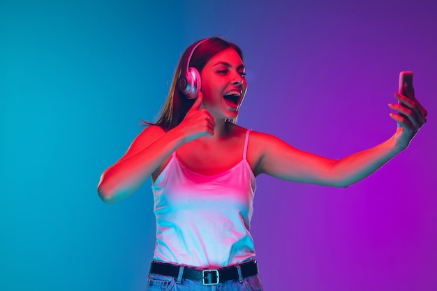 Selfie, écouter de la musique au casque. Portrait de jeune femme caucasienne sur fond dégradé à la lumière du néon. Beau modèle. Concept d'émotions humaines, d'expression faciale, de ventes, d'annonces. Espace de copie.