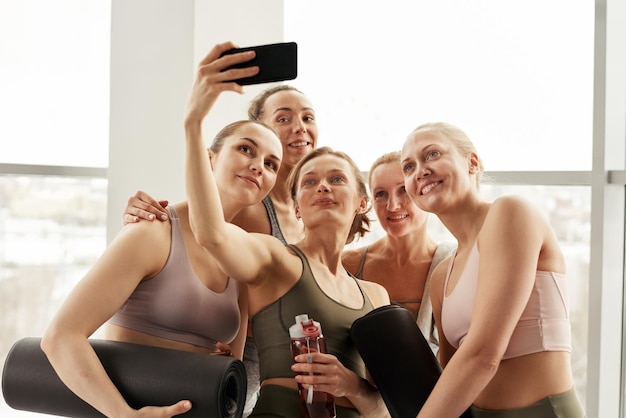 Selfie de groupe de femmes de fitness dans des soutiens-gorge de sport posant avec des tapis de yoga contre la fenêtre en studio