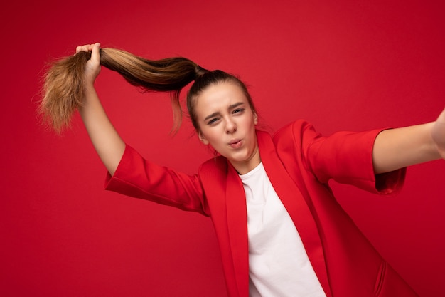 Selfie photo en gros plan d'une jolie petite adolescente brune souriante et souriante portant une veste rouge à la mode et un t-shirt blanc pour une maquette debout isolée sur un mur de fond rouge regardant la caméra