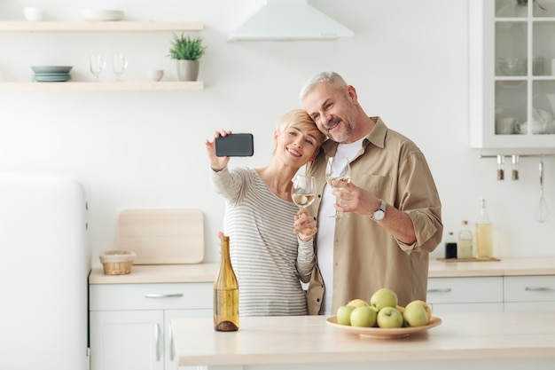 Selfie pour les vacances, application pour téléphone, appel vidéo à la famille pendant covid-19. Femme et homme d'âge moyen joyeux avec du vin dans un intérieur de cuisine confortable, prend une photo sur un smartphone avec écran vide, espace libre