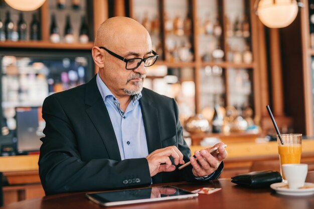 Senior barbu en costume à l'aide d'un téléphone intelligent pour lire ou écrire un message tout en étant assis dans la cafétéria.