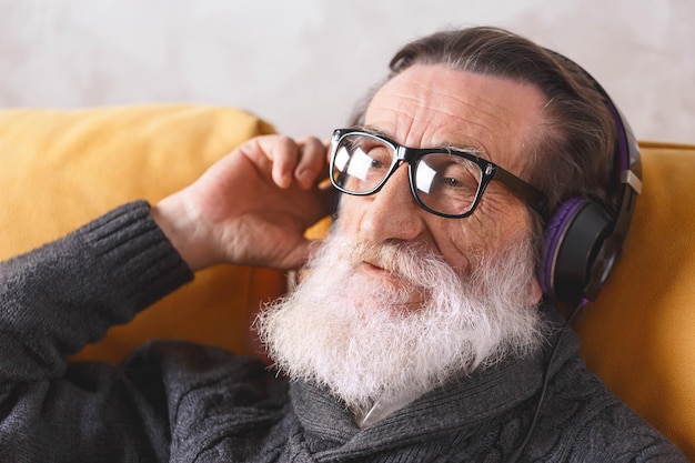 Senior bel homme barbu à lunettes portant un pull gris assis dans des écouteurs sur un canapé jaune dans son salon lumineux et écoutant sa musique préférée concept de communication de technologie moderne