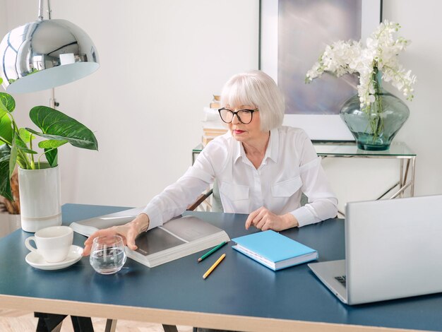 Senior belle femme aux cheveux gris en blouse blanche de l'eau potable pendant le travail au bureau