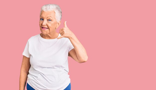 Senior belle femme aux yeux bleus et aux cheveux gris portant un t-shirt blanc décontracté souriant faisant un geste de téléphone avec la main et les doigts comme parler au téléphone communiquer des concepts