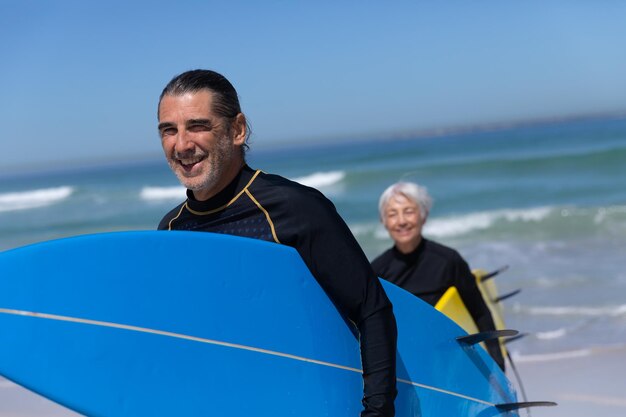 Senior Caucasian couple profitant du temps à la plage par une journée ensoleillée, marchant et tenant des planches de surf avec la mer en arrière-plan