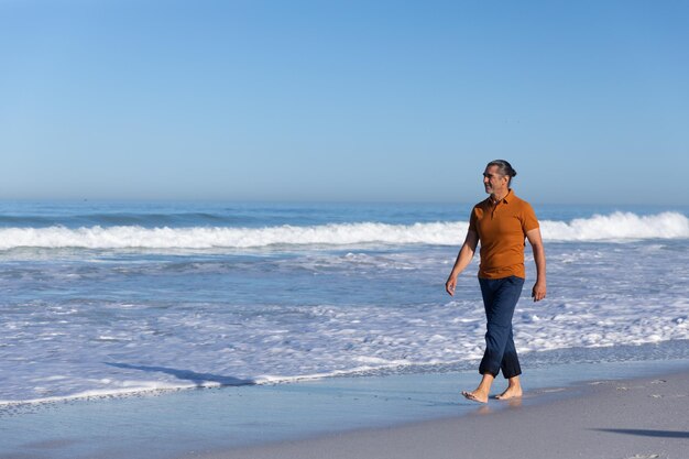Senior Caucasian man profitant du temps à la plage par une journée ensoleillée, marchant pieds nus seul avec la mer en arrière-plan