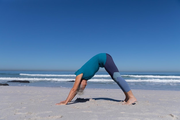 Senior Caucasian woman profitant du temps à la plage par une journée ensoleillée, assis sur le sable et pratiquant le yoga avec la mer en arrière-plan