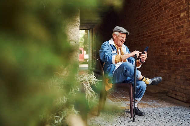 Un senior élégant dans des vêtements à la mode et dans des lunettes est assis sur la chaise et utilise le téléphone.