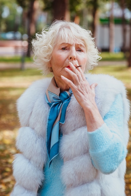 Senior élégant élégant femme à la mode aux cheveux gris en manteau de fourrure cigarette fumer en plein air