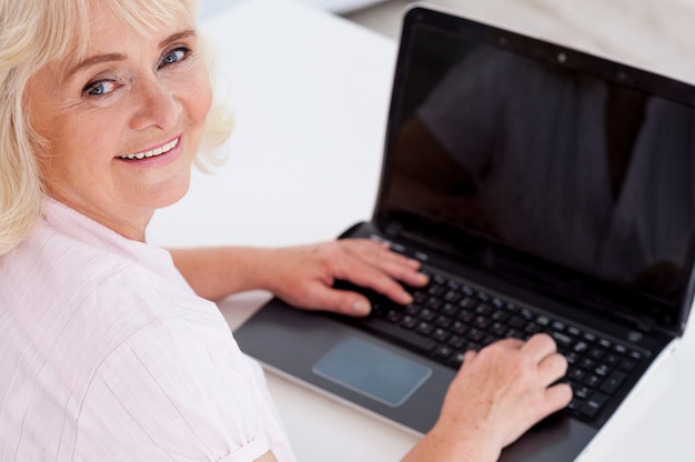 Senior de l'ère numérique. Vue de dessus d'une femme âgée joyeuse regardant la caméra et souriant tout en travaillant sur un ordinateur portable