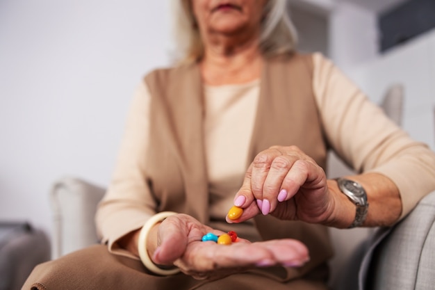 Senior femme assise à la maison dans sa chaise et tenant la main pleine de pilules et de vitamines. Mise au point sélective à portée de main.