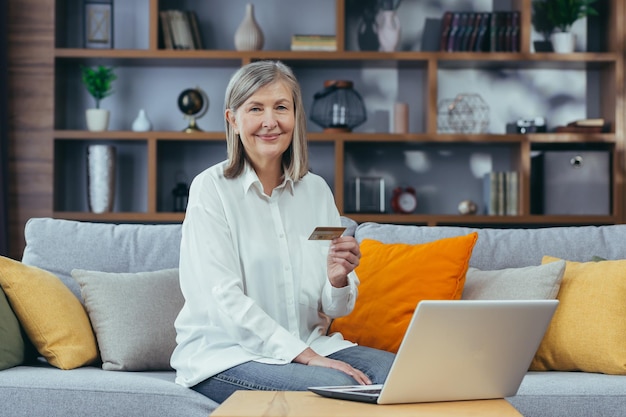 Senior femme aux cheveux gris shopping en ligne portrait de grand-mère avec ordinateur portable à la maison tenant une carte de crédit bancaire regardant la caméra et souriant