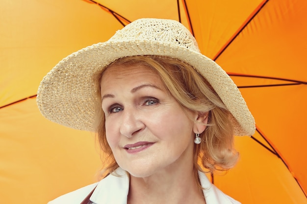 Senior femme blanche de 62 ans aux cheveux blonds et chapeau sur la tête est debout sous un parapluie orange.