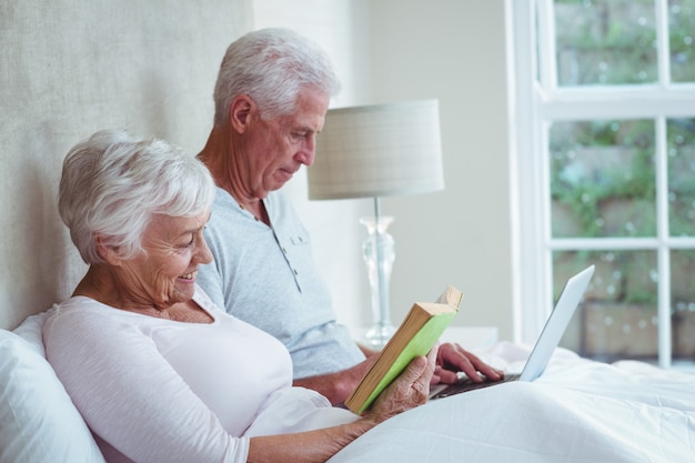 Senior femme lisant un livre tout en utilisant un ordinateur portable