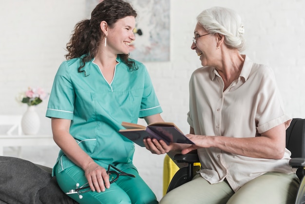 Photo senior femme tenant le livre assis sur le fauteuil roulant se moquer avec belle infirmière