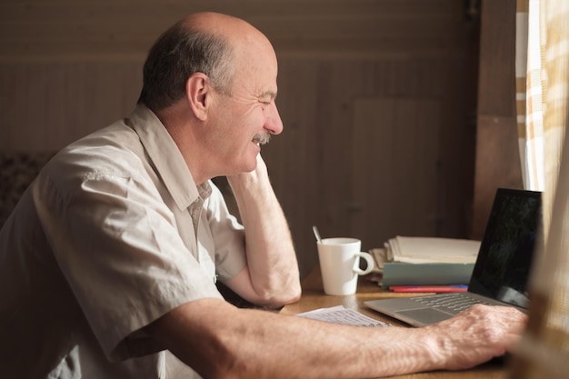 Senior hispanic man sitting near window and looking on photo at latop screen