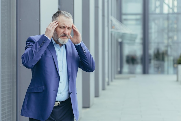 Senior homme d'affaires aux cheveux gris malade à l'extérieur de l'immeuble de bureaux homme tenant les mains sur la tête ayant de graves