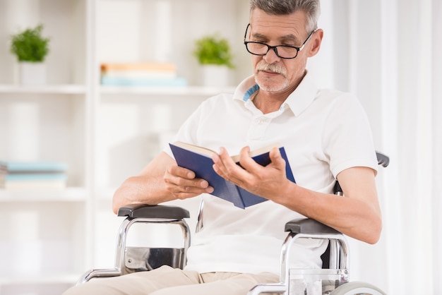 Senior homme assis en fauteuil roulant et en lisant un livre à la maison.