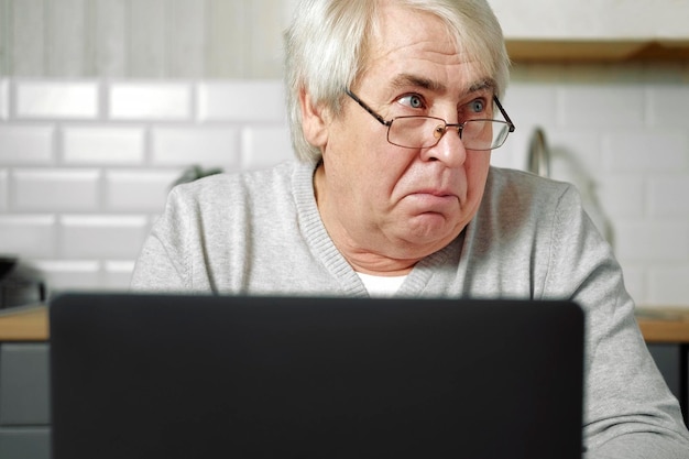 Senior homme aux cheveux gris portant des lunettes assis devant un ordinateur portable et faisant face au poisson avec les lèvres Geste fou et comique Drôle Vieux grand-père avec une expression faciale idiote et ludique grimaçant en train de s'amuser