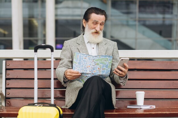 Senior homme à barbe grise est assis sur un banc avec une valise, il regarde la carte de la ville et le téléphone à l'aéroport