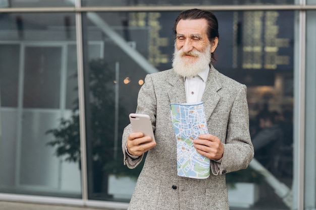 Senior homme à barbe grise à la recherche de smartphone et souriant se promène le long de l'aéroport