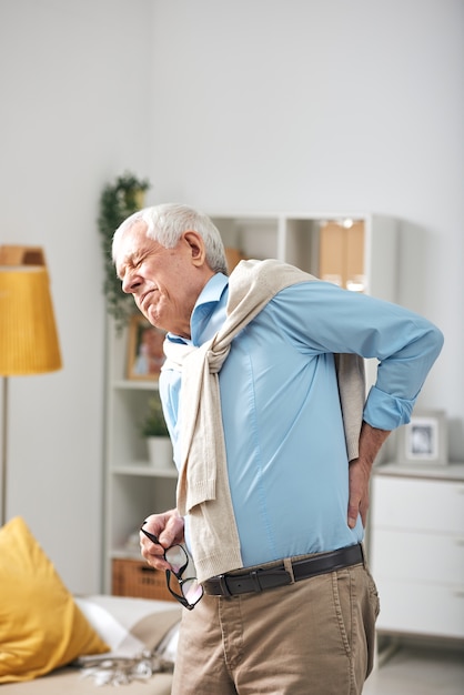 Senior homme debout dans le salon et tenant la main sur le bas du dos tout en ressentant de la douleur