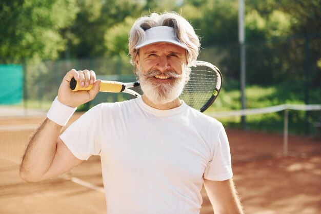 Senior homme élégant moderne avec raquette à l'extérieur sur un court de tennis pendant la journée