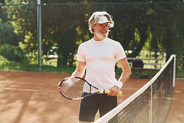 Senior homme élégant moderne avec raquette à l'extérieur sur un court de tennis pendant la journée