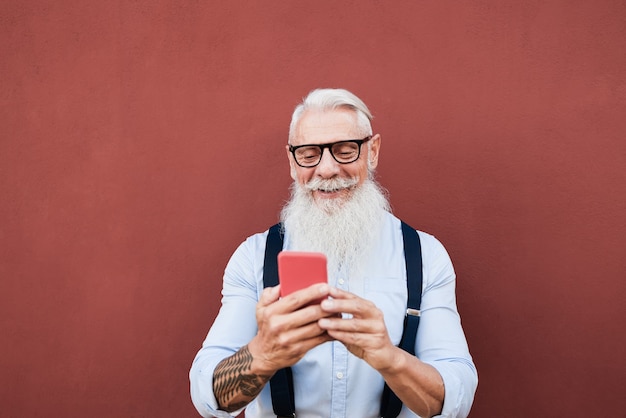Senior homme hipster utilisant un téléphone mobile à l'extérieur avec un mur rouge en arrière-plan - Focus sur le visage