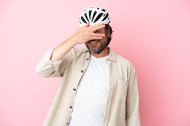 Senior homme hollandais avec casque de vélo isolé sur fond rose couvrant les yeux par les mains et souriant