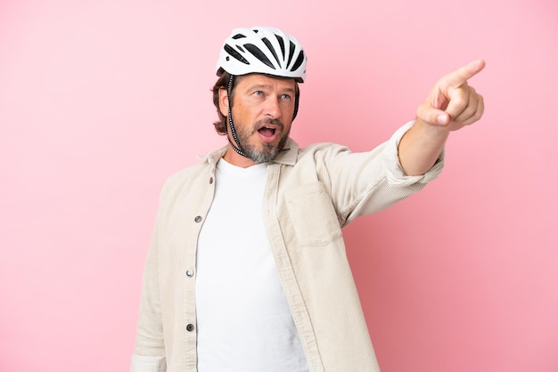 Senior homme hollandais avec casque de vélo isolé sur fond rose pointant vers l'extérieur