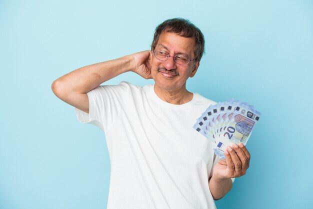 Senior Indian man holding bill isolé sur fond bleu touchant l'arrière de la tête, pensant et faisant un choix.