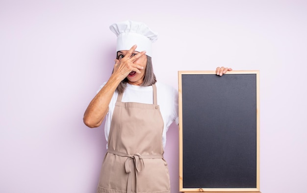 Senior jolie femme regardant choquée effrayée ou terrifiée couvrant le visage avec le chef de la main et le concept de tableau noir