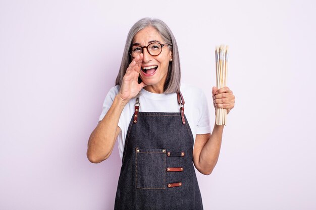 Senior jolie femme se sentant heureuse en donnant un grand cri avec les mains à côté de la bouche artiste avec des pinceaux