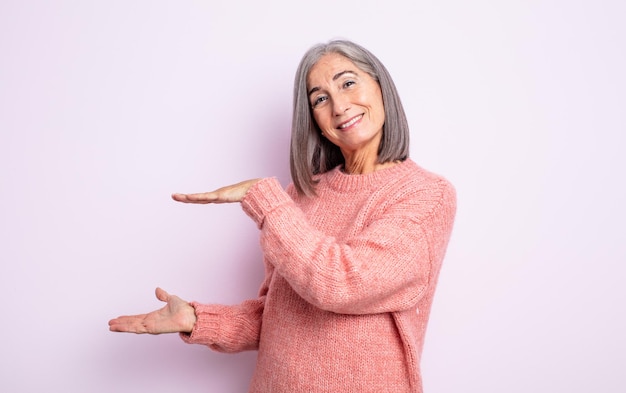 Senior jolie femme tenant un objet avec les deux mains sur l'espace de copie latéral, montrant, offrant ou annonçant un objet