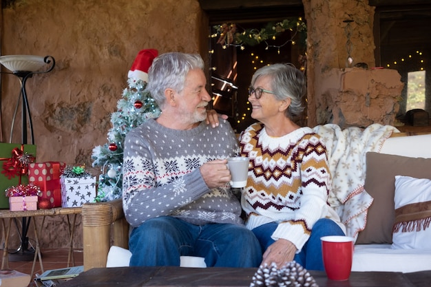 Senior smiling couple sitting on sofa at home au moment de Noël se détendre et se regarder. Arbre de Noël, cadeaux et décorations sur fond