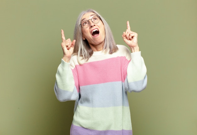 Senior woman choqué, étonné et bouche ouverte, pointant vers le haut avec les deux mains pour copier l'espace