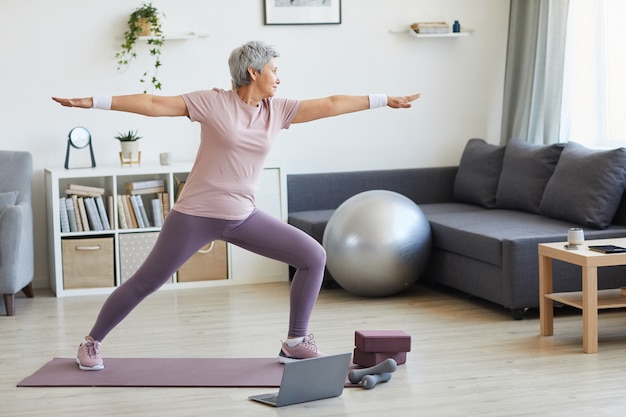 Senior woman debout sur un tapis d'exercice pendant l'entraînement sportif dans le salon à la maison