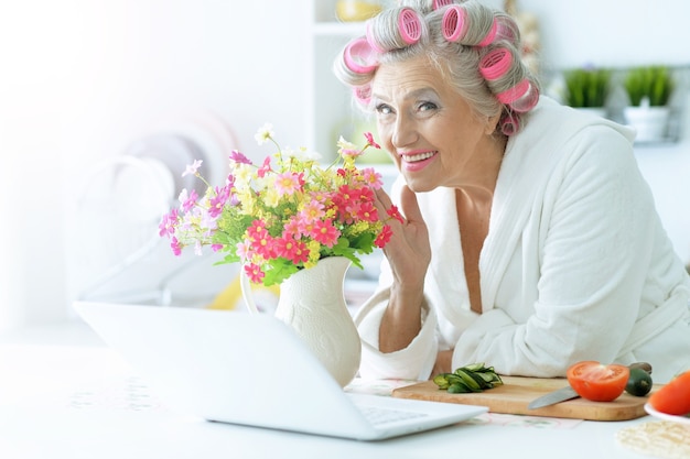 Senior woman in hair rollers à cuisine avec ordinateur portable et légumes