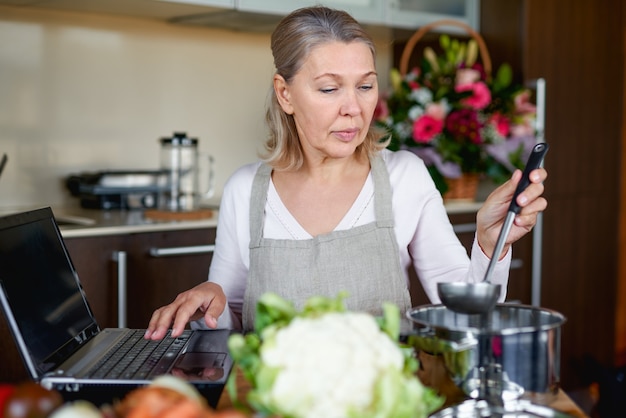 Senior woman in tablier à la cuisine prépare la nourriture et utilise un ordinateur portable