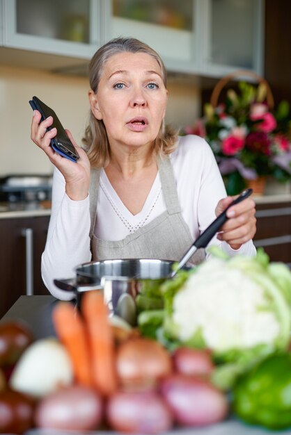 Senior woman in tablier à la cuisine, préparer la nourriture et tenant un téléphone intelligent