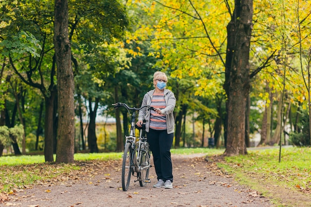 Senior woman marche dans le parc avec un vélo dans un masque médical de protection