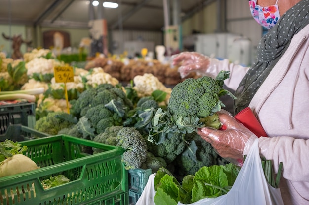 Senior woman sélectionnant le brocoli au marché fermier local. Choix de légumes frais et crus. Femme avec des gants de protection et un masque