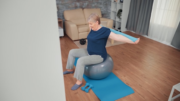 Senior woman sitting on balance ball formation avec bande de résistance. Personne âgée s'entraînant à la maison pour un mode de vie sain, entraînement physique pour personnes âgées en appartement, activité et soins de santé