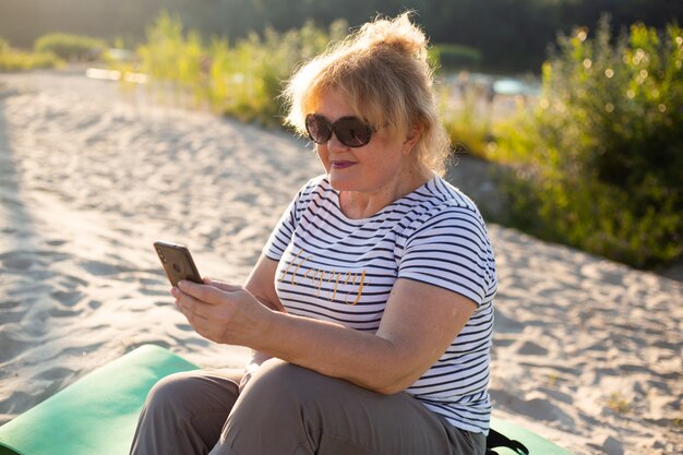 Senior woman sitting on a sand and using smartphone in summer beach