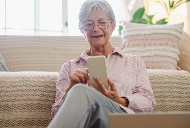 Senior woman sitting on the floor at home tout en lisant un message sur téléphone mobile dame âgée appréciant la technologie et la communication