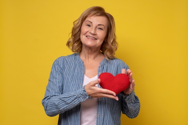 Senior woman smiling montrant coeur rouge sur mur jaune