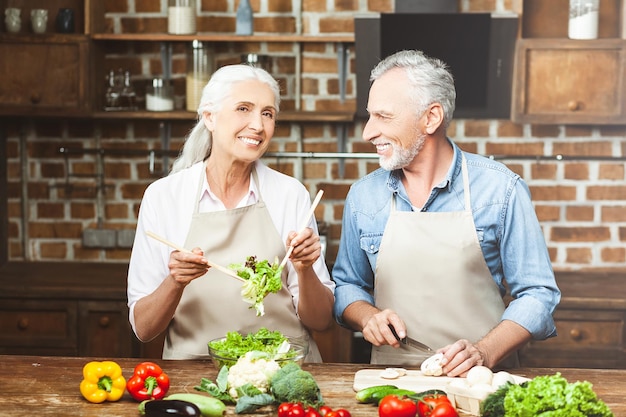 Senior woman tossing salad et regardant la caméra avec un homme coupant des légumes
