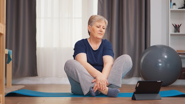 Senior woman warming épaule regardant tutoriel en ligne assis sur un tapis de yoga. Apprentissage et étude en ligne, mode de vie sain et actif, entraînement sportif pour personnes âgées, entraînement à domicile, bien-être et exercice en salle