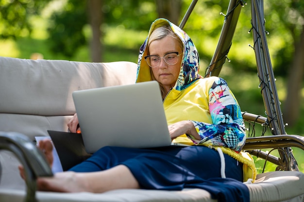 Senior woman with laptop and documents working in garden on rocking couch green home office concept